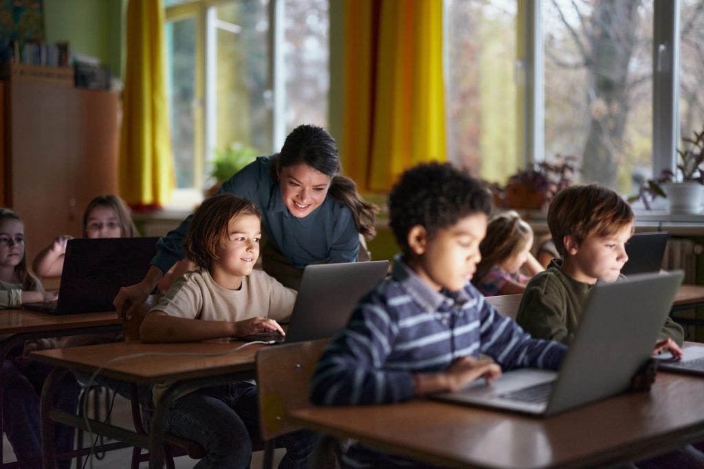 A classroom scene where a diverse group of elementary-aged students work on laptops at their desks. A smiling teacher leans over to assist one student, creating an engaging and supportive learning environment. Sunlight streams through large windows with yellow curtains in the background.